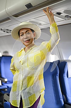 Portrait of 50s woman tourist standing at airplane aisle and smiling to camera. Retirement, travel and summer vacation