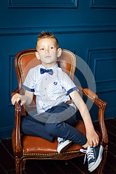 Portrait of 7 years old caucasian boy in blue bow tie and blue sneakers sitting in big brown leather chair. Fashionable good