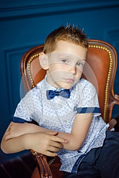 Portrait of 7 years old caucasian boy in blue bow tie sitting in big brown leather chair. Fashionable good looking little boy sit