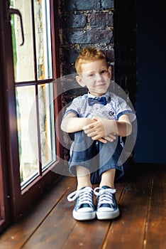 Portrait of 7 years old caucasian boy in blue bow tie with sly smile and his hands on knees. Fashionable good looking little boy