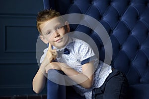 Portrait of 7 years old caucasian boy in blue bow tie with sly smile and his hands near face. Fashionable good looking little boy