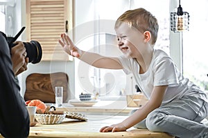 Portrait of aborable boy model posing in front of photographer. Kid sitting on table in bright kitchen room