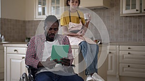 Portrait of absorbed African American disabled man in wheelchair watching at tablet as teenage girl drinking tea coffee