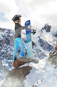 Portrait of active female snowboarder in funny hat standing on top of the mountain background