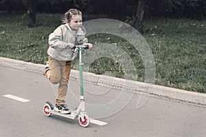 Portrait of active little toddler girl riding scooter on road in park outdoors on summer day. Seasonal child activity