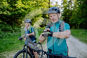 Portrait of active senior couple riding bicycles at summer park, looking at sports smartwatch, checking their