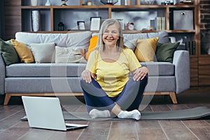 Portrait of active senior woman sitting at home on lotus floor and looking at camera, happy grandmother doing yoga online using