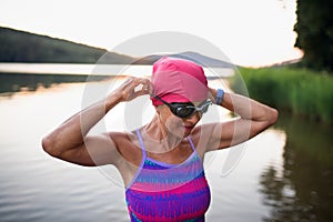 Portrait of active senior woman swimmer standing and putting on goggles outdoors by lake.