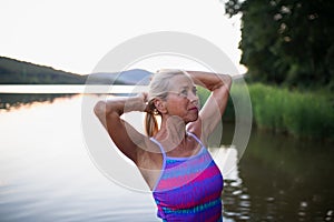 Portrait of active senior woman swimmer standing and stretching outdoors in lake.