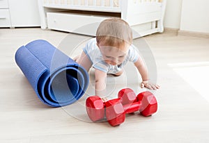Portrait of active toddler boy crawling on floor towards yoga mat and red dumbbells. Concept of children sport