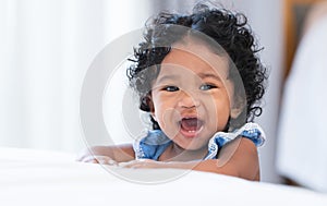 Portrait of adorable African 7 months old newborn baby girl with black curly hair standing near bed at home, looking at camera,