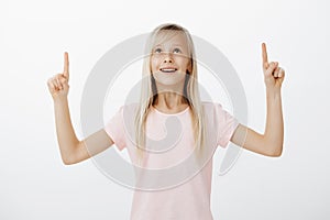 Portrait of adorable amazed child with blond hair in pink t-shirt, raising index fingers, pointing and looking up with