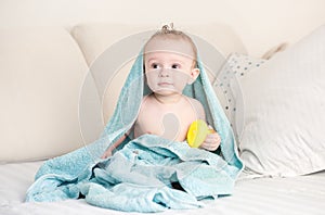 Portrait of adorable baby boy covered in blue towel sitting on bed with yellow rubber duck