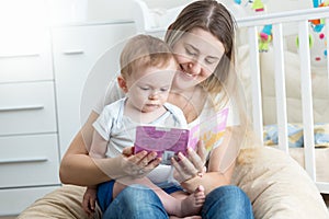 Portrait of adorable baby boy readig book with smiling young mother in bedroom