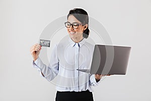 Portrait of adorable businesswoman wearing eyeglasses holding silver laptop and credit card in the office, isolated over white