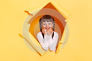 Portrait of adorable charming little girl with braids wearing casual shirt looking through torn hole in yellow paper, looking at