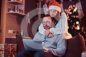 Portrait of an adorable couple - charming woman in Santa hat hugging her man and using a laptop.