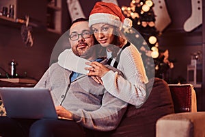 Portrait of an adorable couple with laptop - charming woman in Santa hat hugging her man and looking at camera.