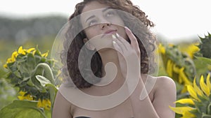 Portrait of a adorable curly girl in black bra posing and touching her hair standing in the sunflower field. Bright