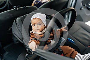 Portrait of adorable infant baby boy sitting on front seat with buckled security belt in car, looking at camera. Cute 6