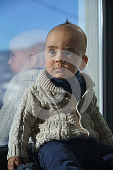 Portrait of an adorable kid in beige sweater sitting on window sill
