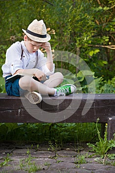 Portrait of adorable kid boy in a straw hat sitting on a wooden bench and reading book in the park. Child reading in the garden on