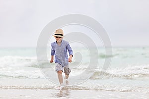 Portrait of adorable kid boy in straw hat and sun glasses running on ocean beach and looking at the waves. Vacations by the sea.