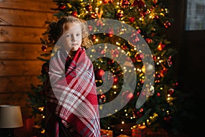 Portrait of adorable little blonde curly child girl wrapped in plaid looking at camera standing by decorated Christmas