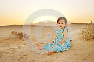 Portrait of adorable little girl on beach vacation weared beautiful blue dress. Baby sitting on sand in sunset time.