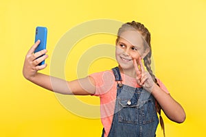 Portrait of adorable little girl in denim overalls gesturing victory or peace while communicating on mobile phone