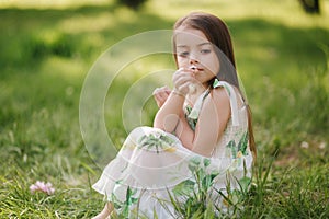 Portrait of adorable little girl sits barefoot on the grass in the park. Happy kid on the fresh air