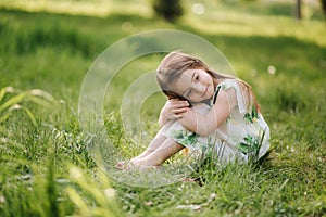 Portrait of adorable little girl sits barefoot on the grass in the park. Happy kid on the fresh air