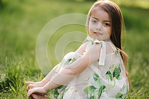 Portrait of adorable little girl sits barefoot on the grass in the park. Happy kid on the fresh air