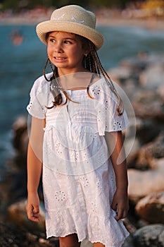 Portrait of adorable little girl in white dress and knitted hat stand on rocks by the sea.