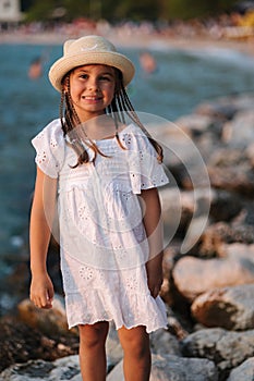 Portrait of adorable little girl in white dress and knitted hat stand on rocks by the sea.