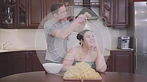 Portrait adorable plump woman sitting in the kitchen at home. The slim blond man hanging noodles on the ears of his fat