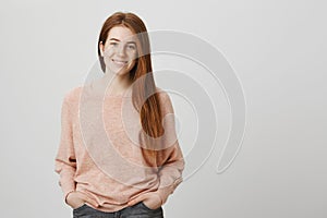 Portrait of adorable redhead caucasian woman standing with hands in pockets over gray background, smiling and expressing