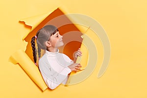 Portrait of adorable smiling little girl with braids wearing casual shirt looking through torn hole in yellow paper, looking away