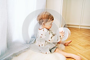portrait of adorable toddler girl sitting on the floor by the window, playing with toy drum