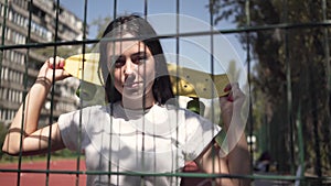 Portrait of adorable woman with a skateboard looking at the camera standing behind the mesh fence outdoors. Concept of