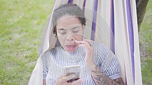 Portrait of adorable young African American woman lying in the hammock, relaxing in the garden, texting on cell phone