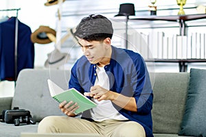 Portrait adult asian man sitting at sofa reading book, look at computer and thinking new project