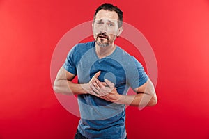 Portrait of adult bearded man 30s having touching his chest having heart attack or suffering from hypertension, isolated over red