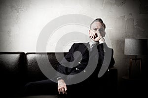 Portrait of adult businessman wearing trendy suit and sitting modern studio on leather sofa against the empty concrete
