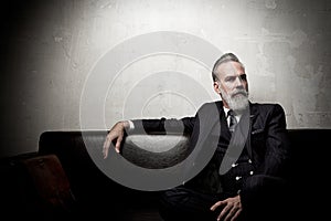Portrait of adult businessman wearing trendy suit and sitting modern studio on leather sofa against the empty concrete