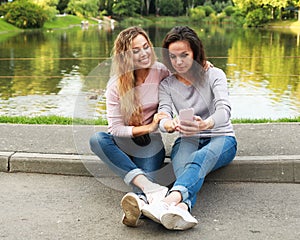 Portrait of Adult caucasian daughter showing on mobile phone to her senior mother outdoor in summer park, near lake