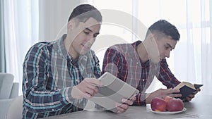 Portrait of adult Caucasian man playing video game on tablet and showing it to his twin brother. Brunette guy reading