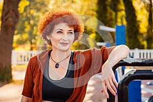 Portrait of adult Caucasian smiling woman with makeup and ginger curly hair posing in sports park. Concept of healthy