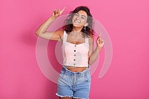 Portrait of adult happy brunette woman pointing up with her index fingers, standing isolated over pink background, expressing