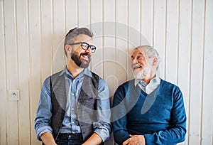 A portrait of adult hipster son and senior father sitting on floor indoors at home.